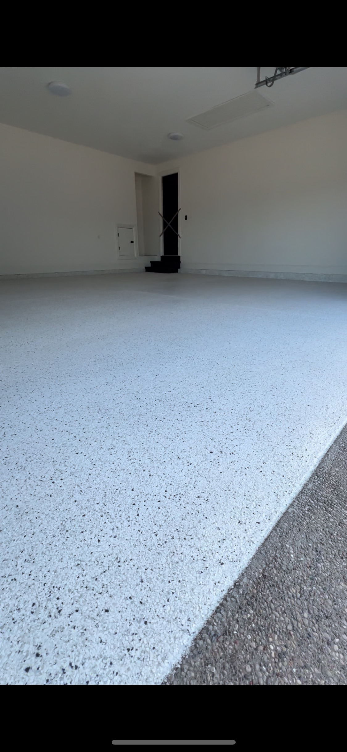 An empty garage with a speckled floor and white walls. A dark doorway is at the far end.