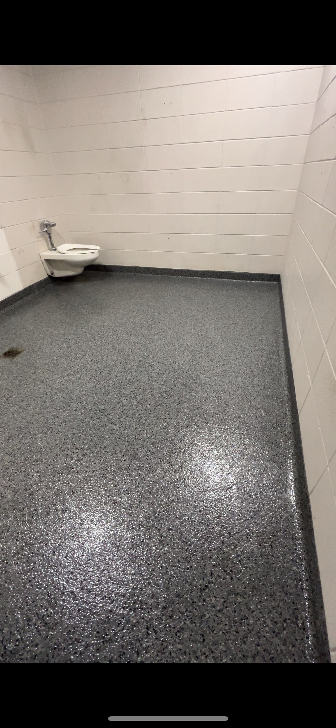 A public restroom with a speckled gray floor and white tiled walls. A toilet is visible on the left.