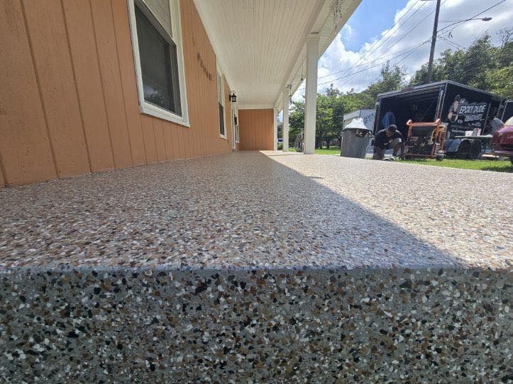 A low-angle view of a speckled, concrete porch with an edge. The porch runs alongside a brown building, under a white awning.