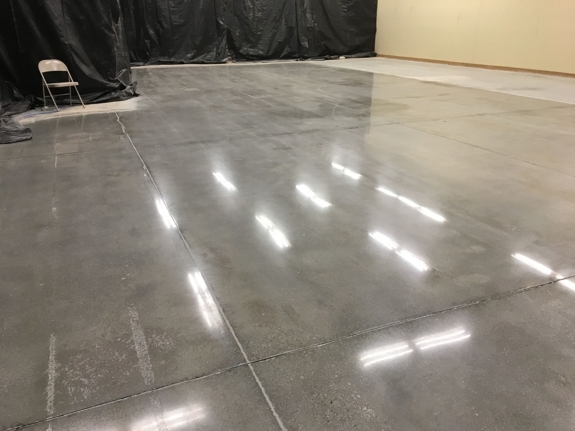 Polished concrete floor in an empty room, reflecting overhead lights. Black plastic covers a wall.