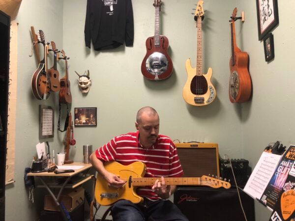 David Dunavant playing the guitar in his studio with guitars hanging on the walls in the background.