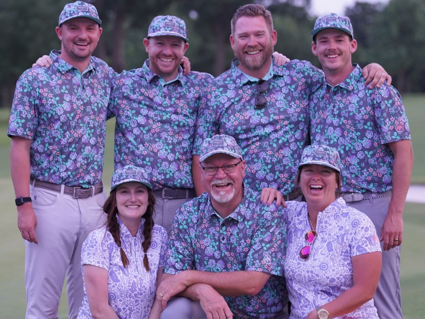 Group of people smiling, wearing matching floral shirts and hats on a golf course.
