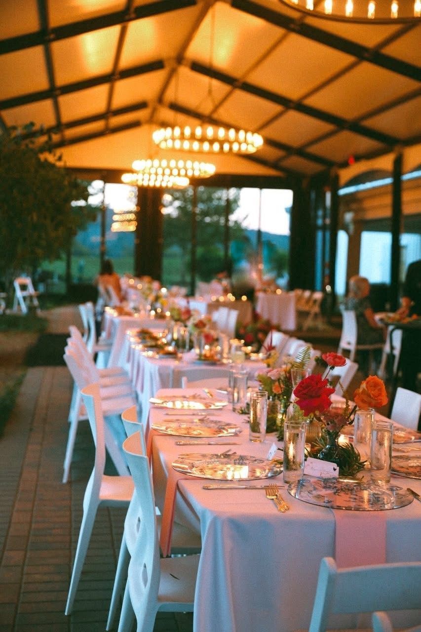 Long dining tables set for a celebration under a canopy with floral centerpieces and white chairs.