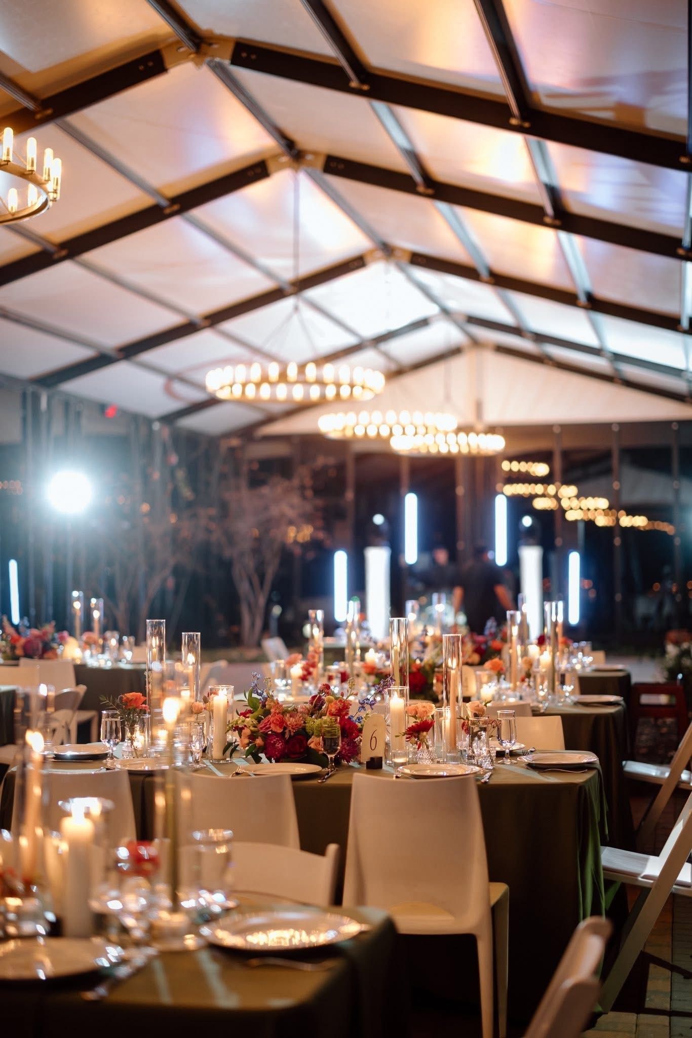 Inside event tent: tables set with candles, flowers, and place settings. Overhead lights and white chairs.