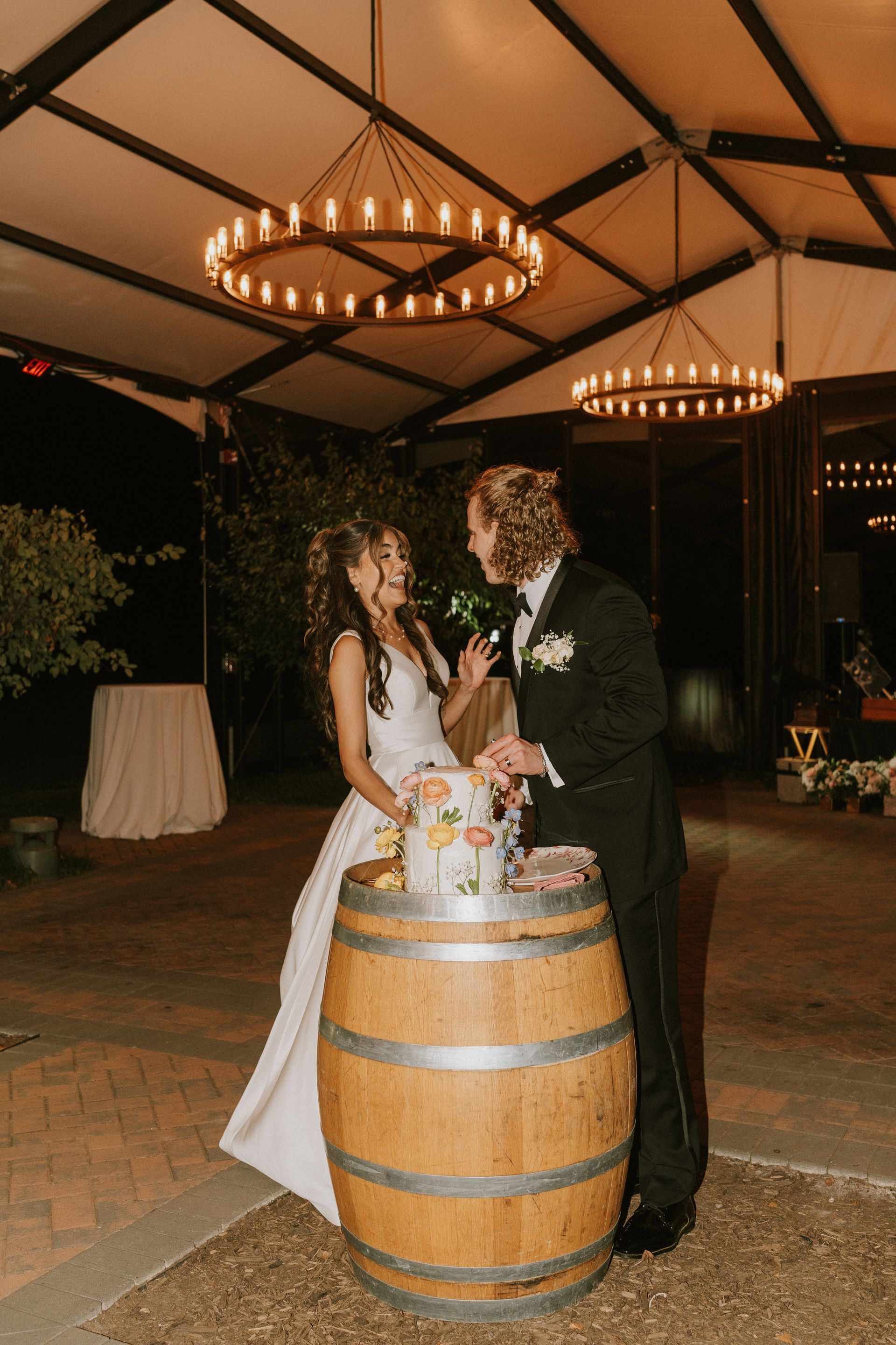 Bride and groom cut wedding cake atop a wooden barrel at night, under chandeliers.