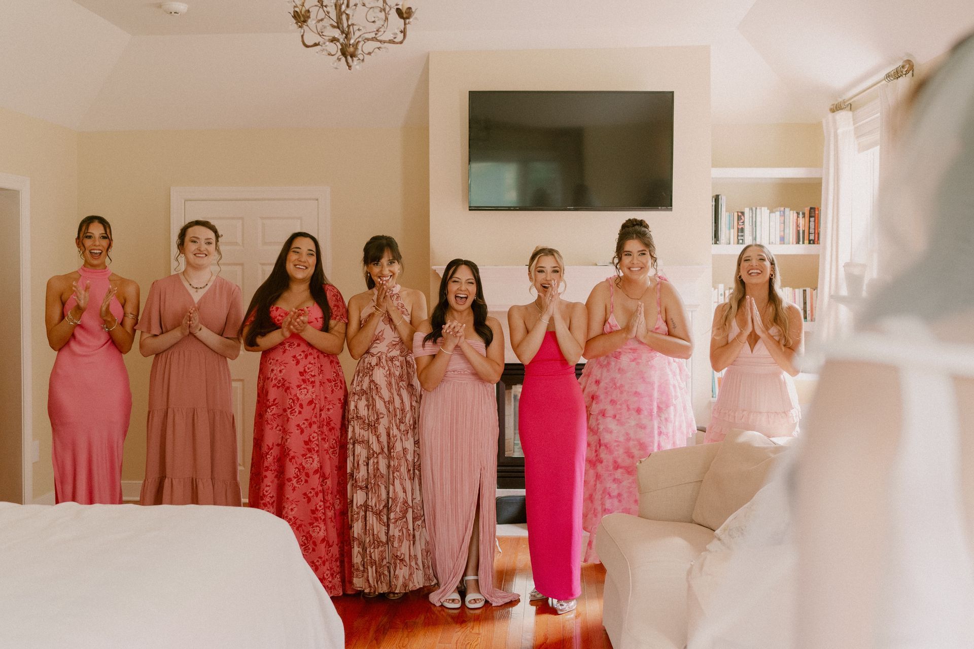 Bridesmaids in pink dresses react with surprise, hands clasped, as they see the bride. Indoor setting.