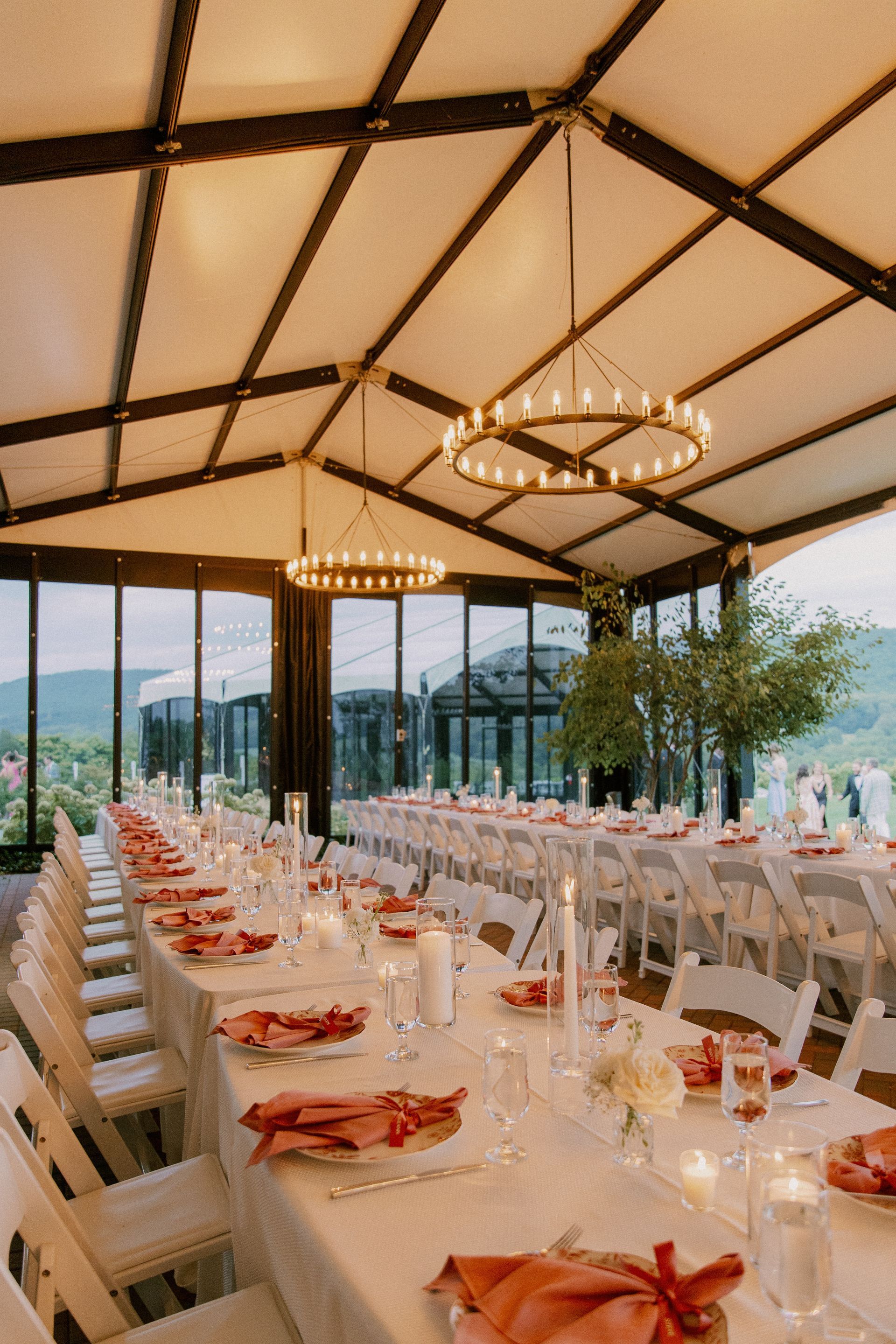 Long banquet tables set for a celebration in a light-filled room with chandeliers and views of mountains.