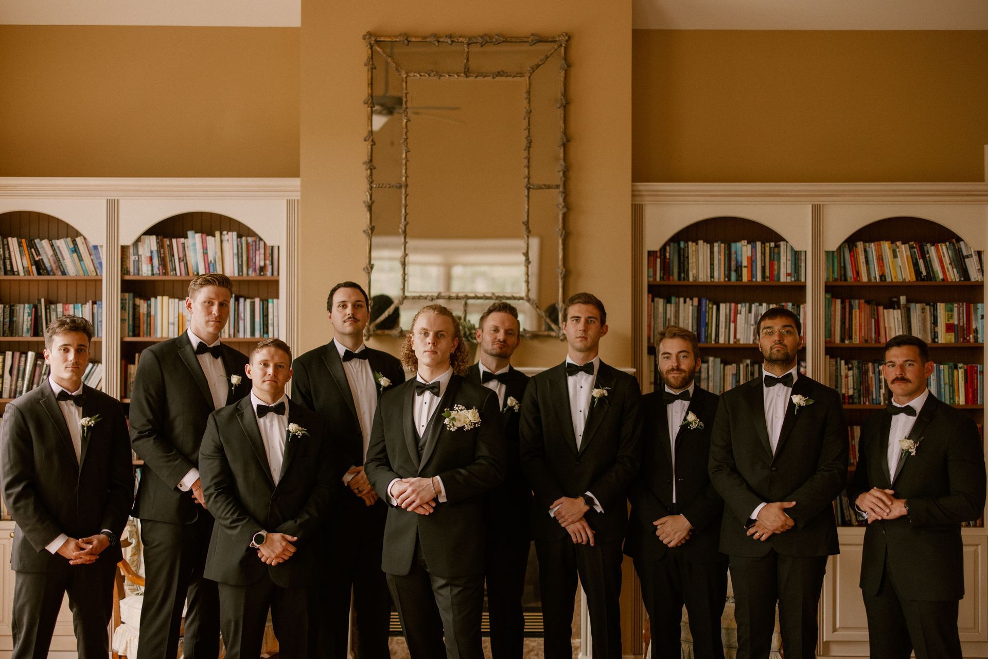 Groomsmen in black tuxedos pose in front of bookshelves and a mirror in a well-lit room.