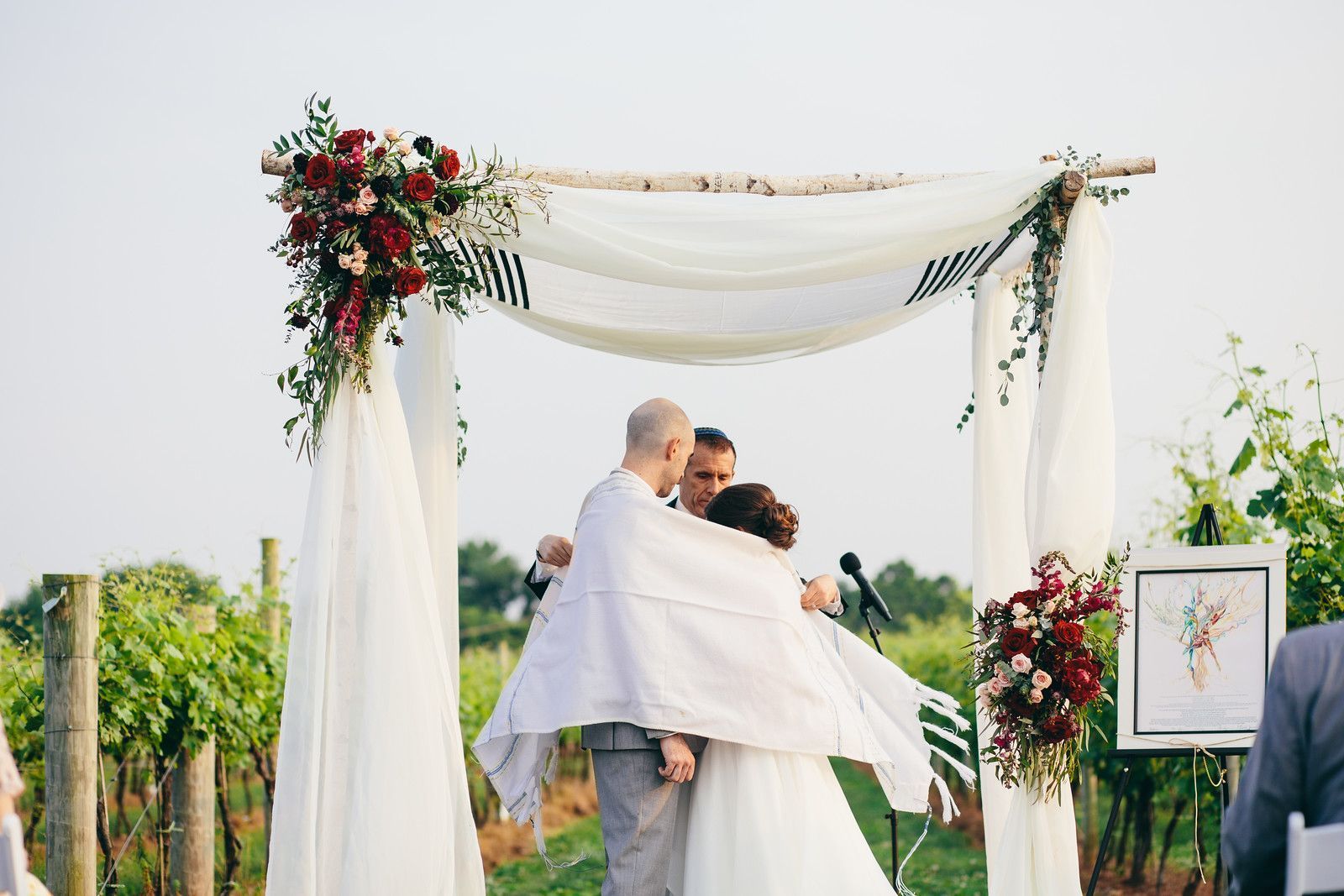 A bride and groom are hugging under an arch at their wedding ceremony.