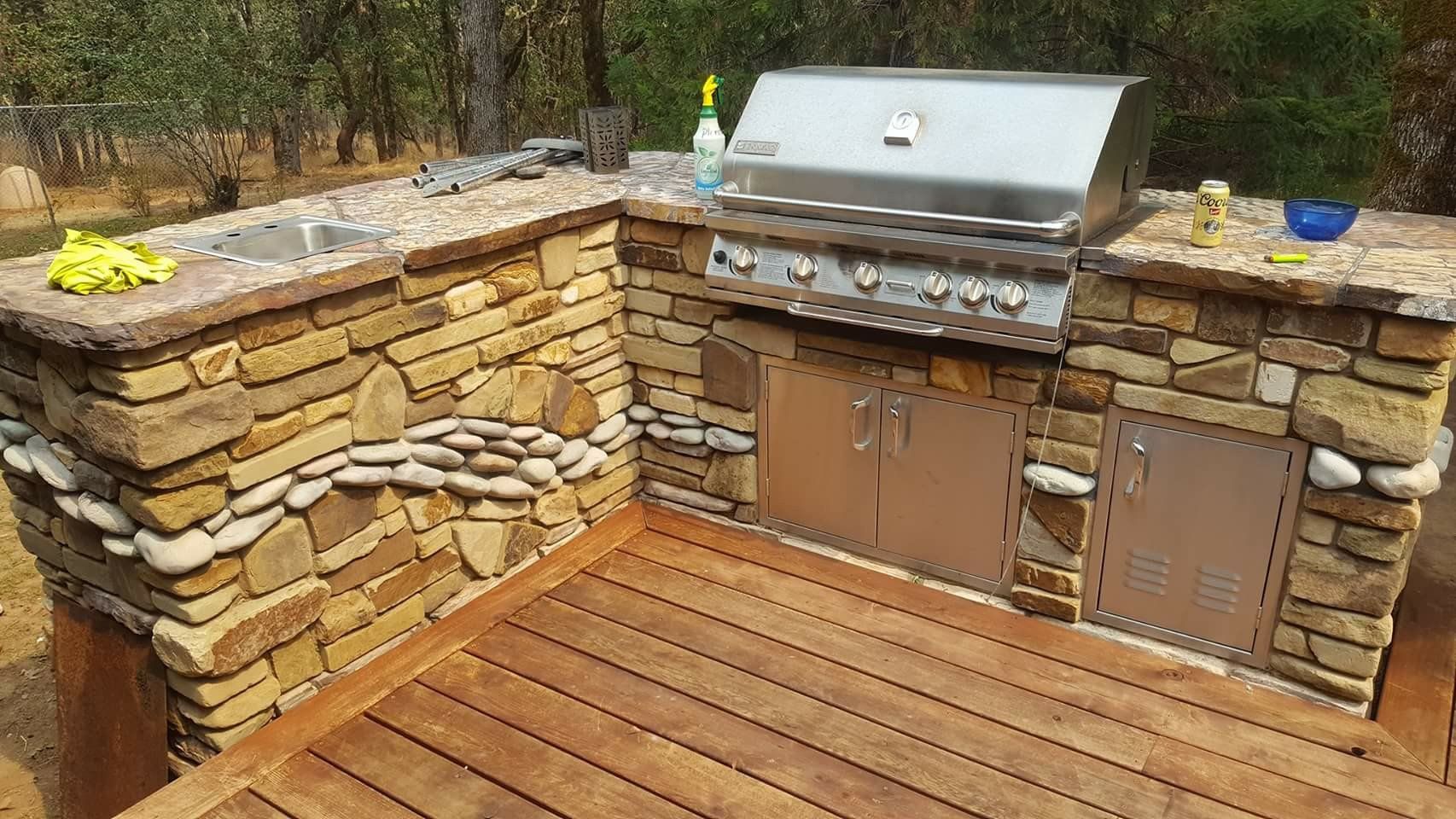 Outdoor stone barbecue grill built into a wooden deck with a stainless steel lid and side cabinets