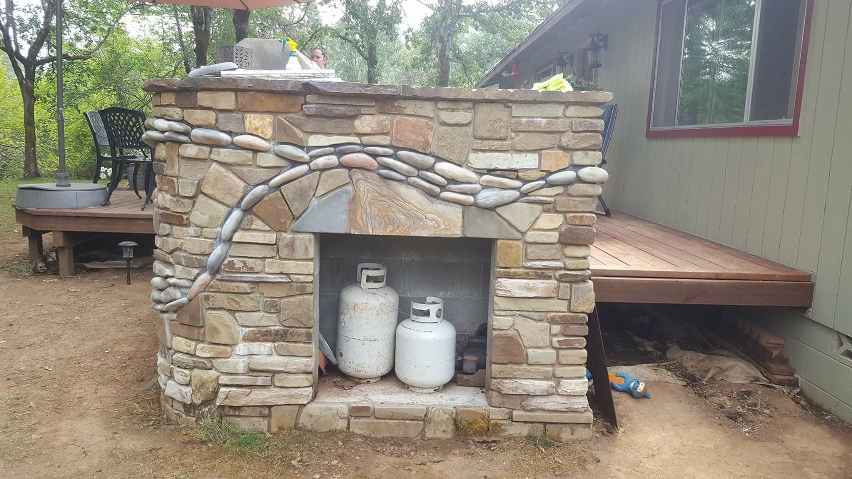 Stone outdoor fireplace with arched stonework and two white propane tanks in a rustic yard