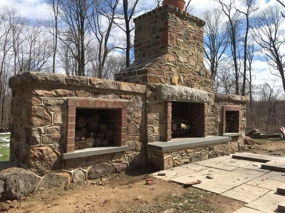 Stone outdoor fireplace with three built-in openings and a tall chimney in a wooded yard