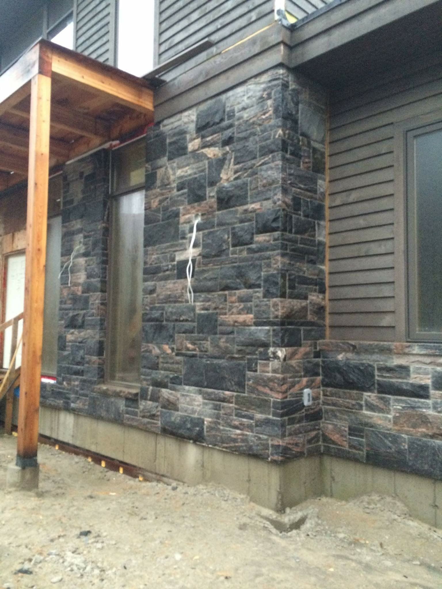 Stone-faced exterior wall with a wooden porch support and concrete foundation on a dirt ground