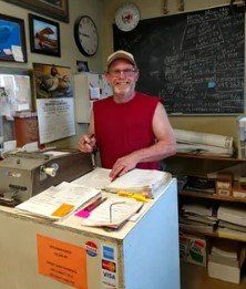 Man standing behind a counter in a cluttered office, smiling with papers and notices around him.