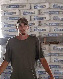 Person standing in front of stacked bags, wearing a cap and dark shirt, indoors.