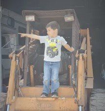 Child standing on a construction loader, arms outstretched, in a garage or workshop.