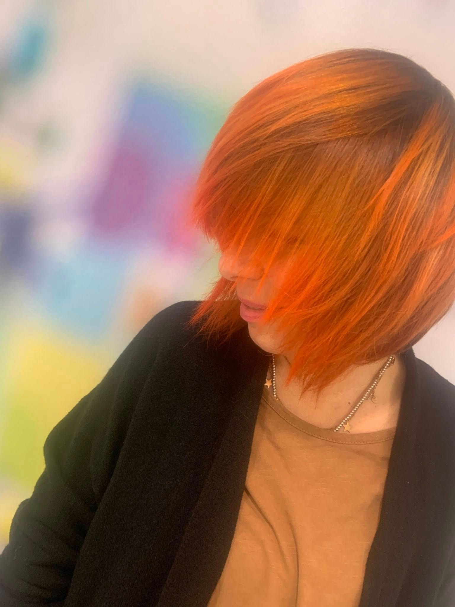 Persona con capelli color arancione acceso, che indossa una giacca nera e una camicia marrone, e guarda in basso.