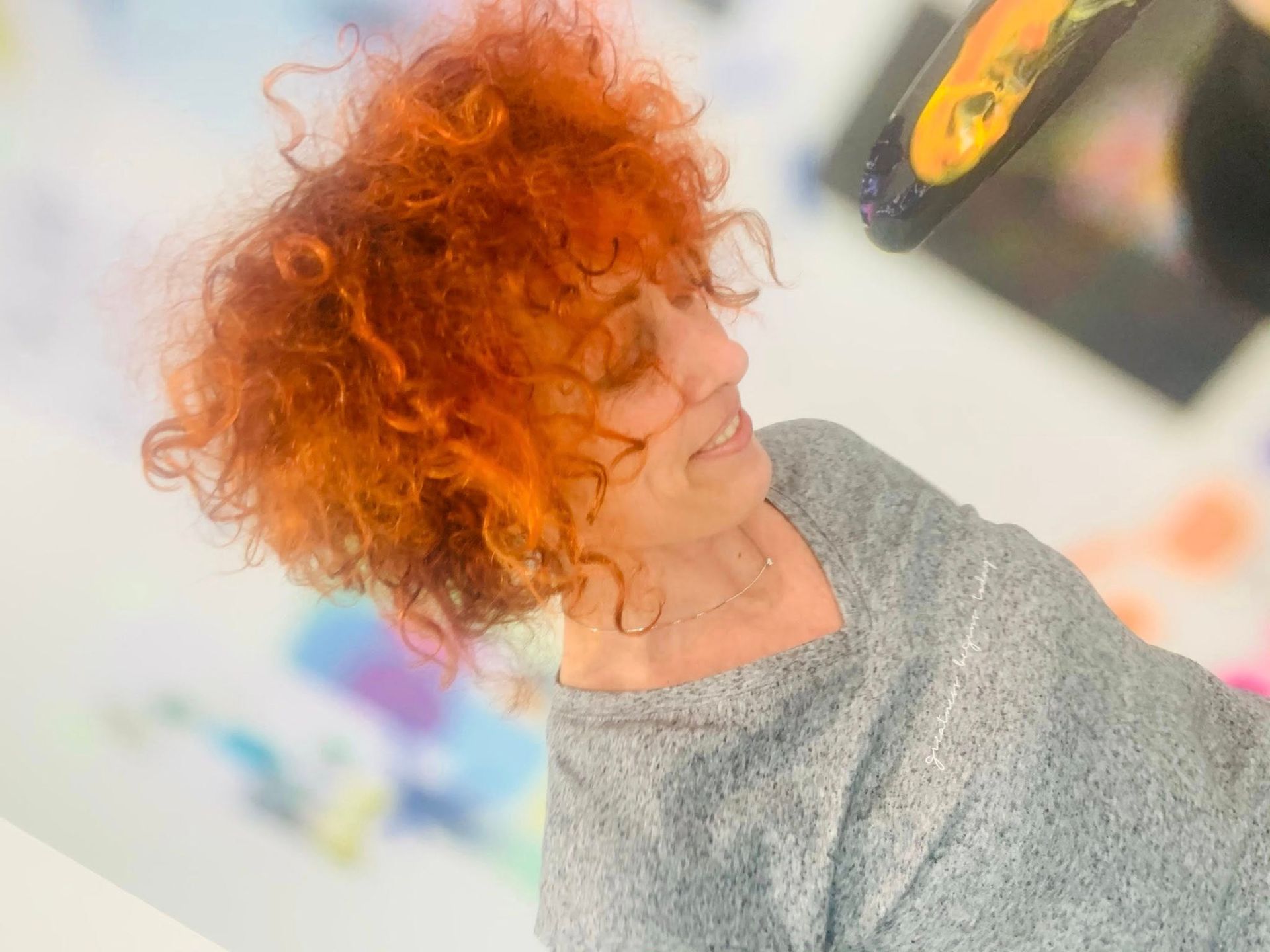 Una donna con i capelli rossi e ricci sorride, guardando in alto. Indossa un maglione grigio ed è in piedi davanti a un'opera d'arte colorata.