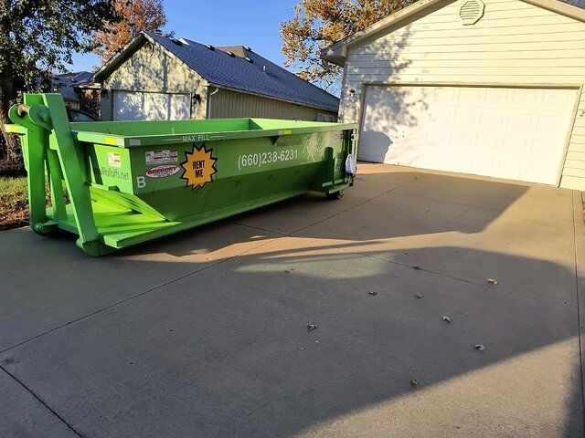 Green dumpster container in a driveway beside a garage, with houses and trees in the background.