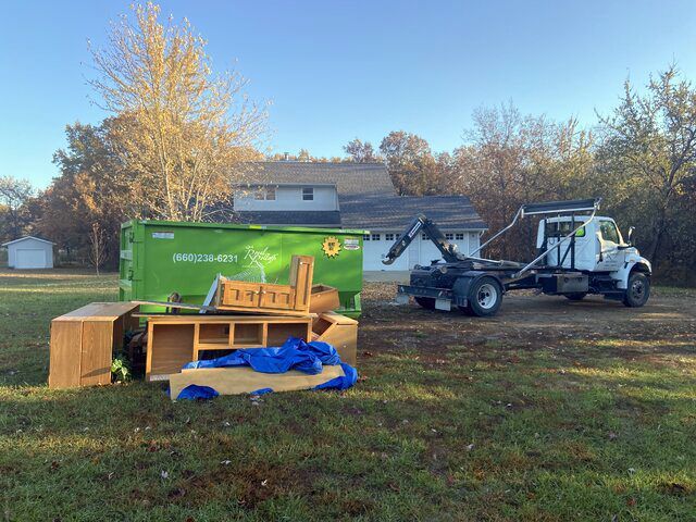 Backyard cleanup with green dumpster, equipment trailer, and construction gear on grass