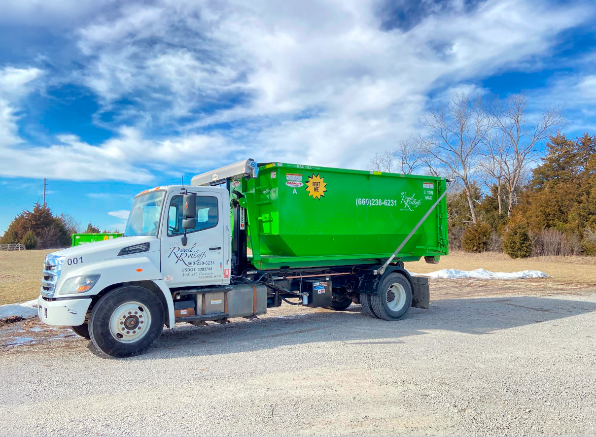 White truck with a green dumpster body parked on a gravel lot under a blue, cloudy sky