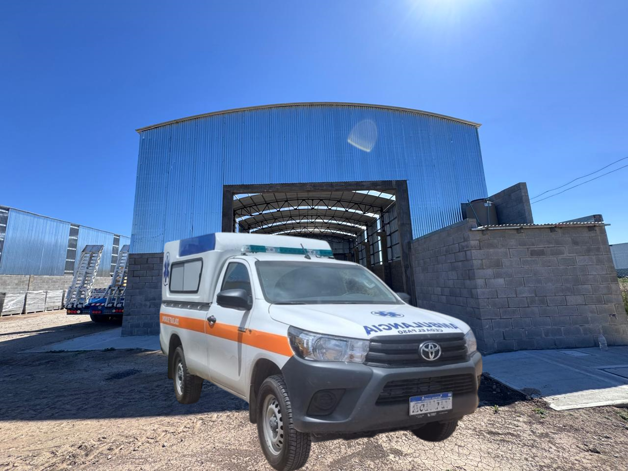 Camión ambulancia blanco frente a un gran edificio metálico bajo un cielo azul.