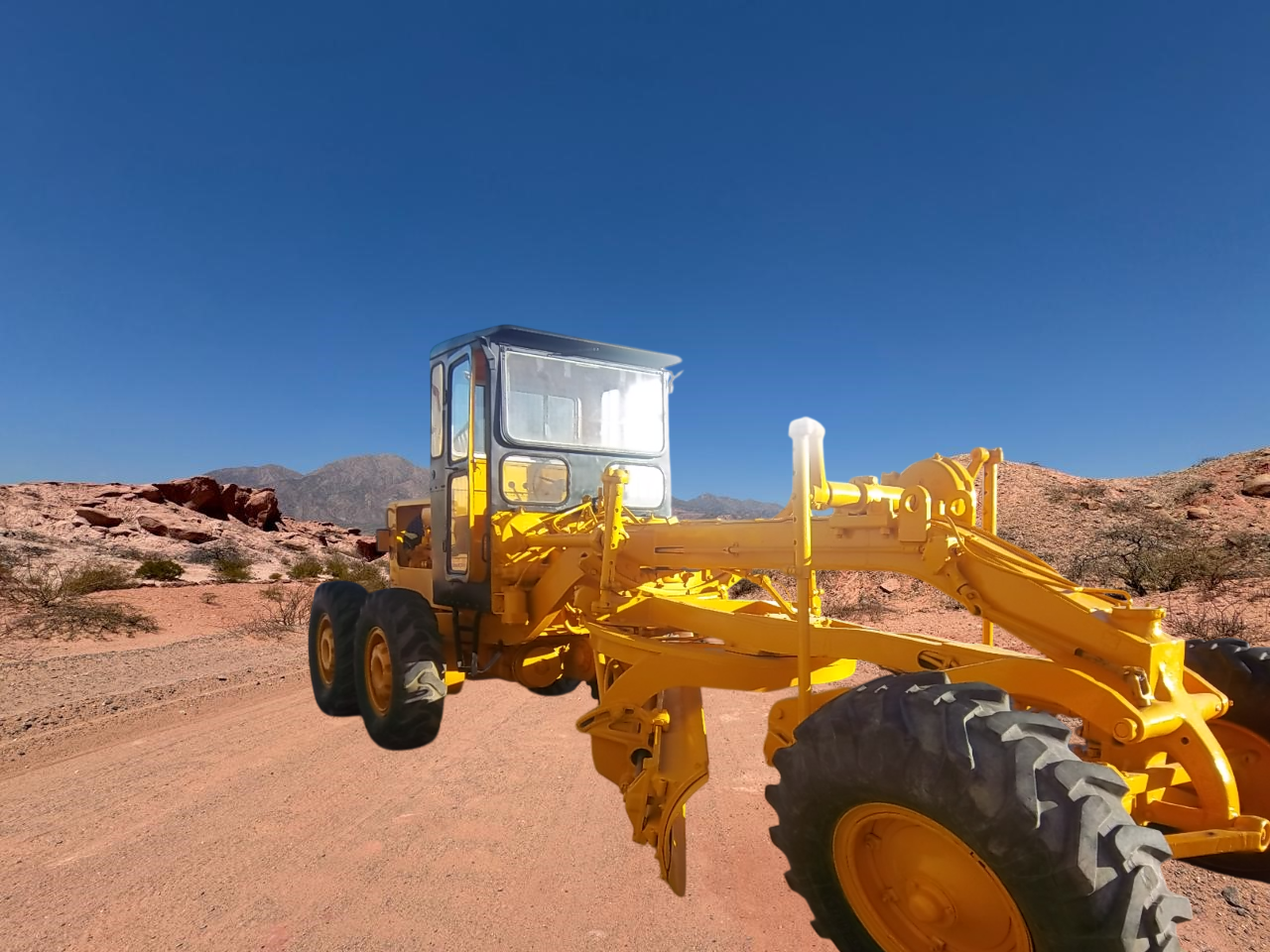 Máquina niveladora amarilla en un camino de tierra en un paisaje desértico con cielo azul.