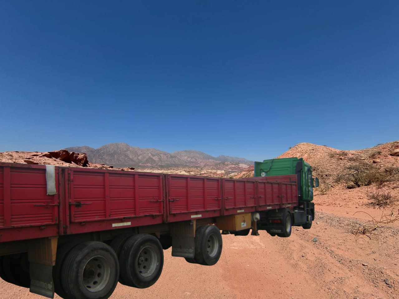 Camión semirremolque rojo y verde en un camino de tierra en un paisaje desértico con montañas y un cielo azul.