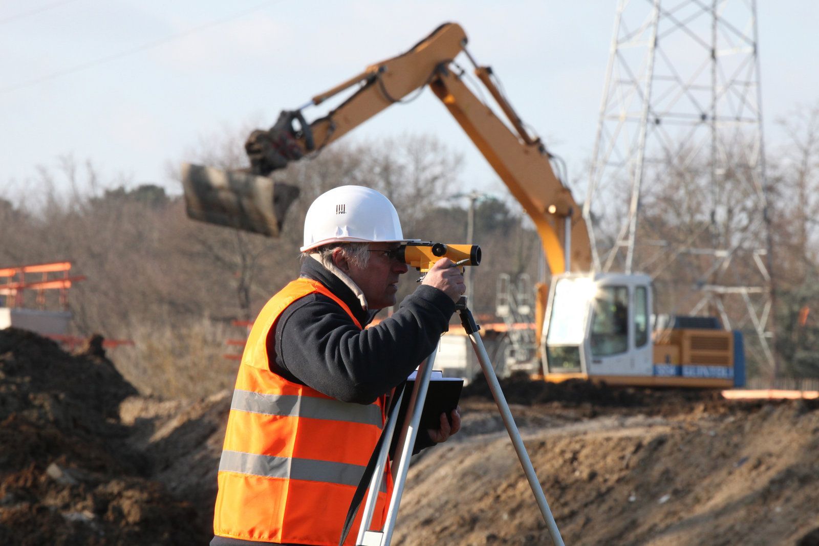 Construction worker in a hard hat and safety vest uses a level. An excavator and power lines are in the background.