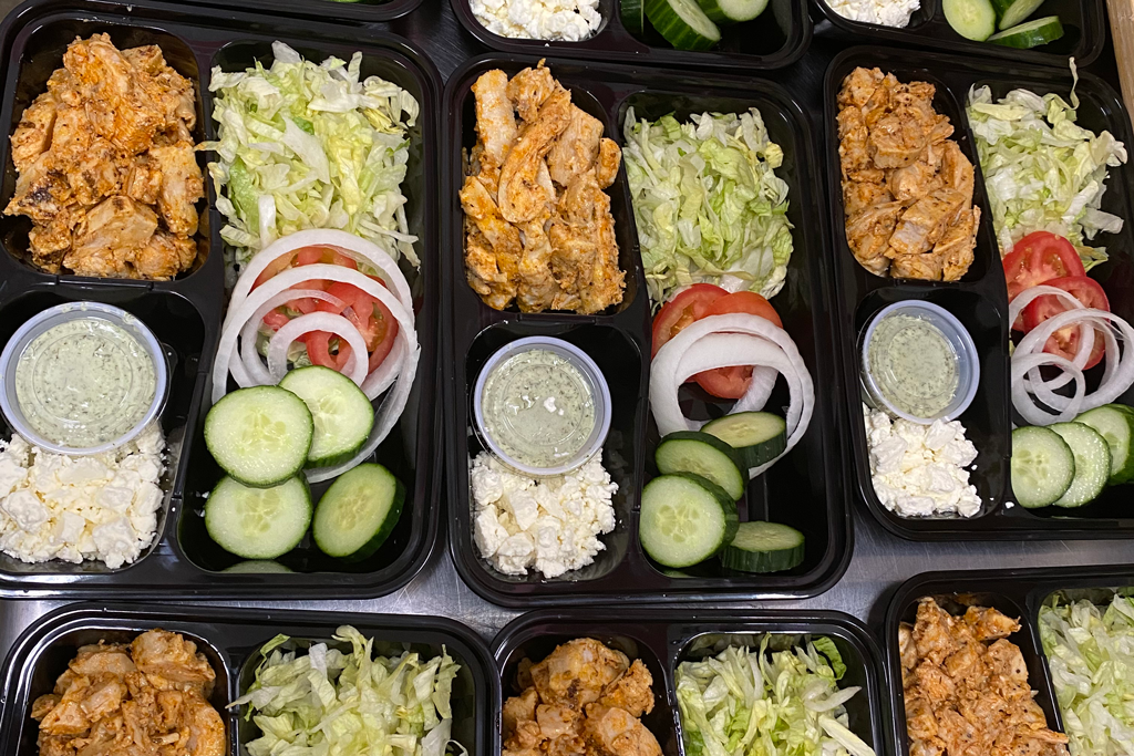 Rows of black meal-prep trays with chicken, rice, and sliced cucumbers, tomatoes, and lettuce.