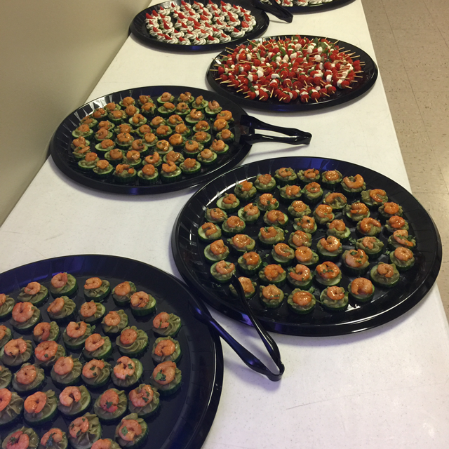Four black trays of bite-sized appetizers arranged on a white tablecloth at a catering event.
