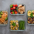 Four meal prep containers with chicken, broccoli, tomatoes, avocado, corn, green beans, and salad.