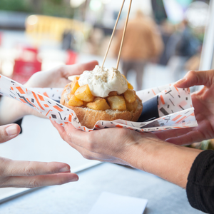 Hands passing a paper tray with a dessert topped with whipped cream and caramel sauce at a table