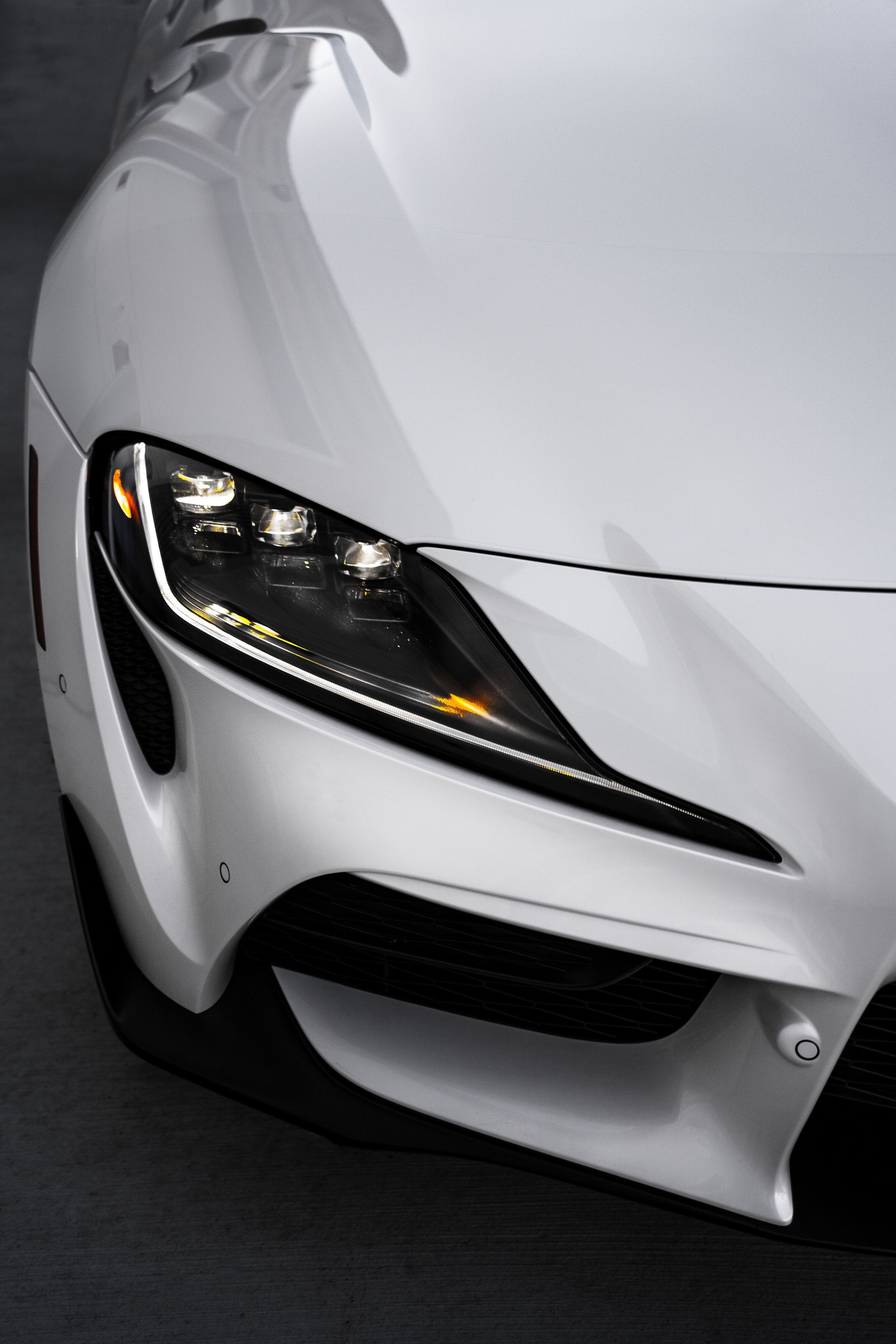 Close-up of a white sports car’s front-left headlight and bumper on a dark road