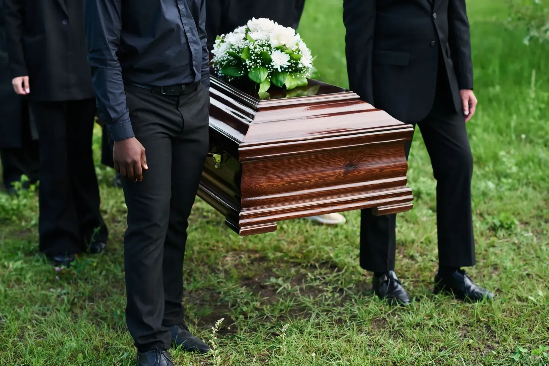 Pallbearers carrying a polished wooden coffin with white flowers across grass at a funeral