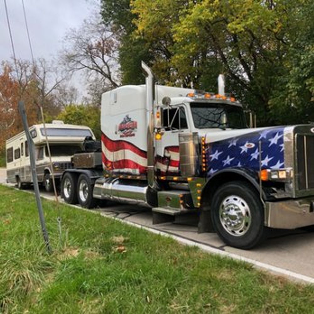 Semi-truck towing a beige camper trailer. Truck has American flag design on the front and side.