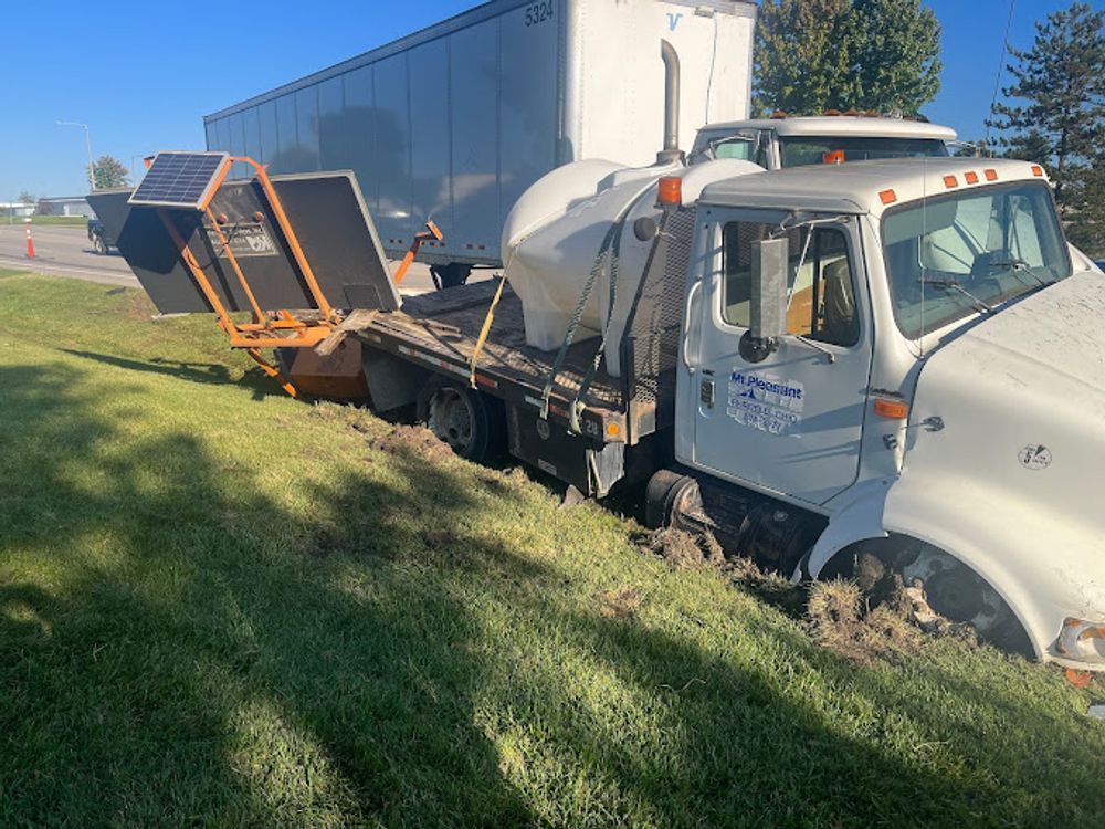 White truck stuck in grass beside a road, with a trailer in the background.