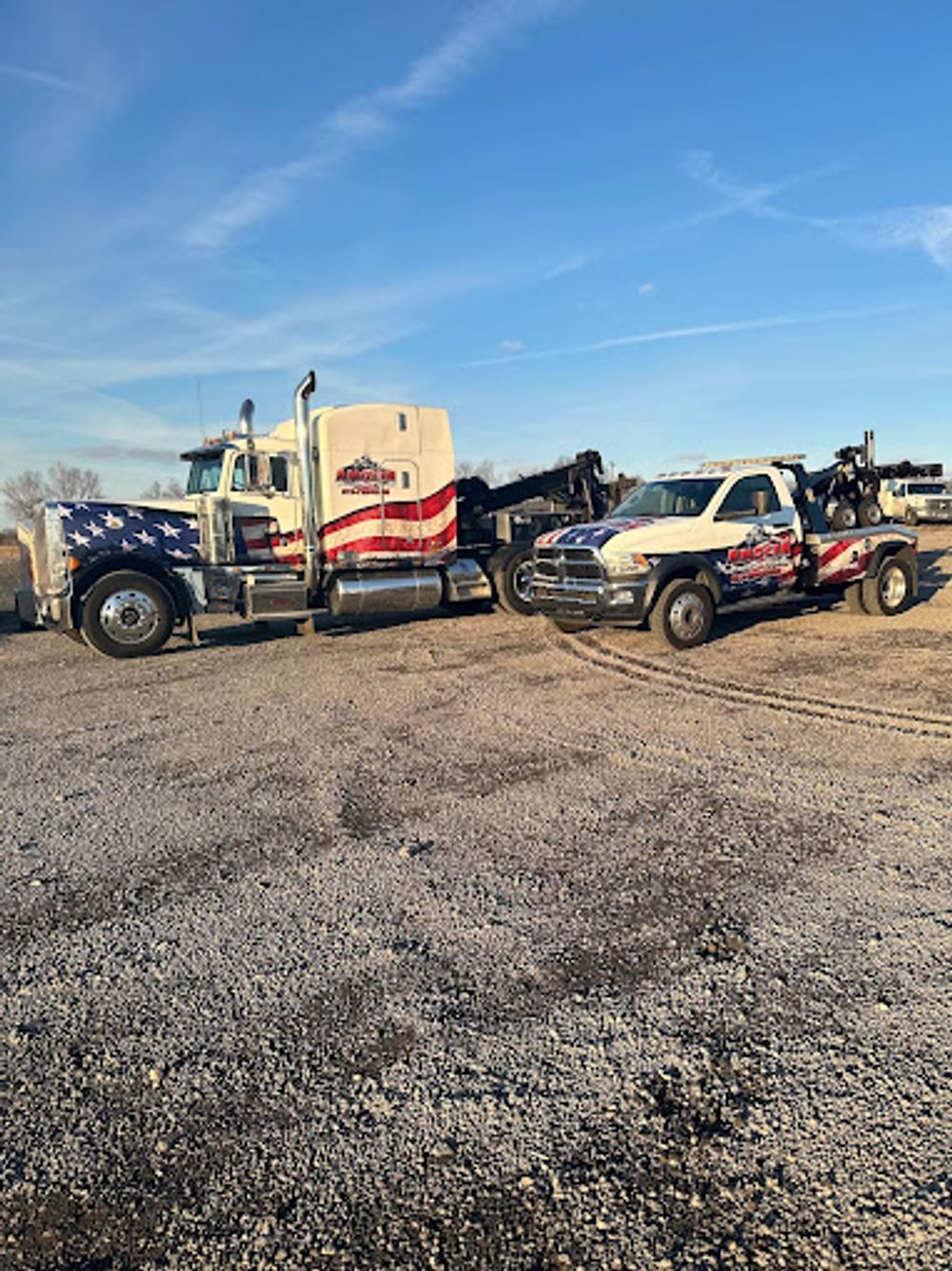 Two tow trucks, one large and one smaller, parked on a gravel lot under a blue sky. Both are painted with American flag themes.