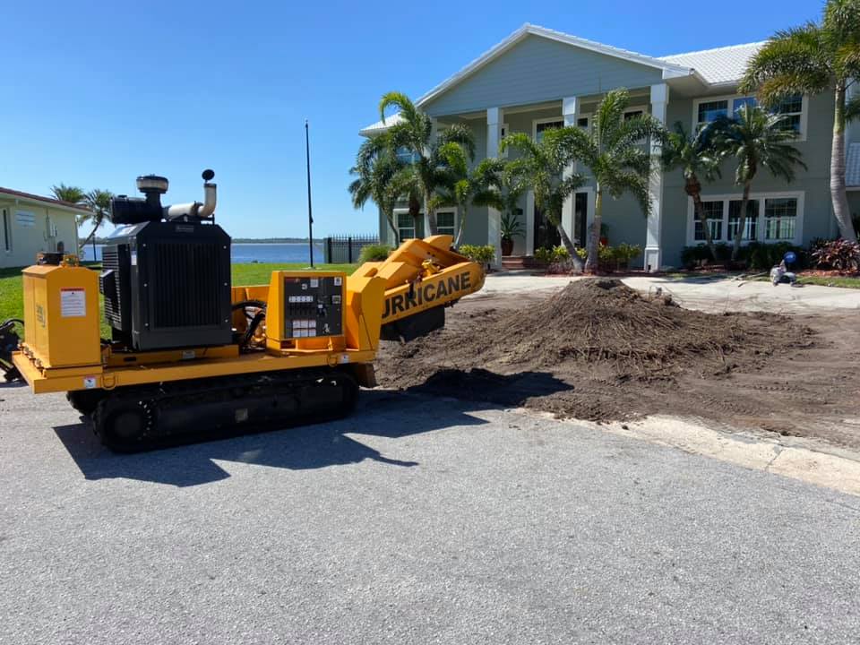 Yellow stump grinder on a driveway, grinding a stump near a waterfront house.