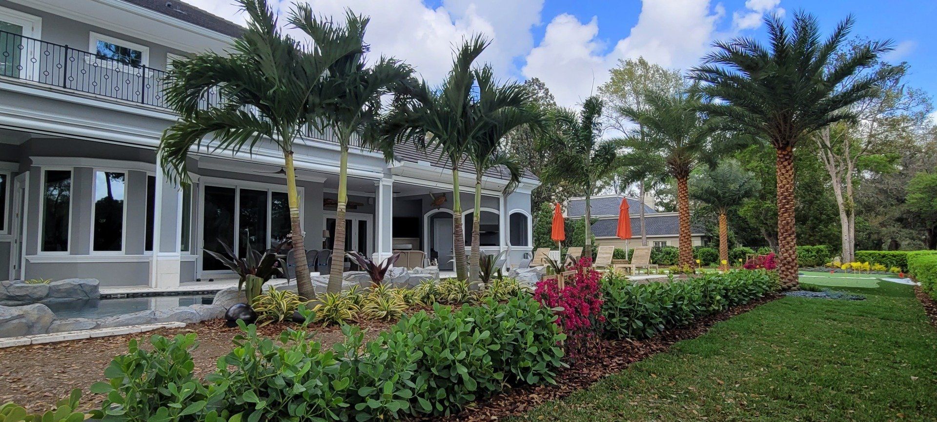 A luxurious white house with palm trees, green lawn, and vibrant garden beds on a sunny day.