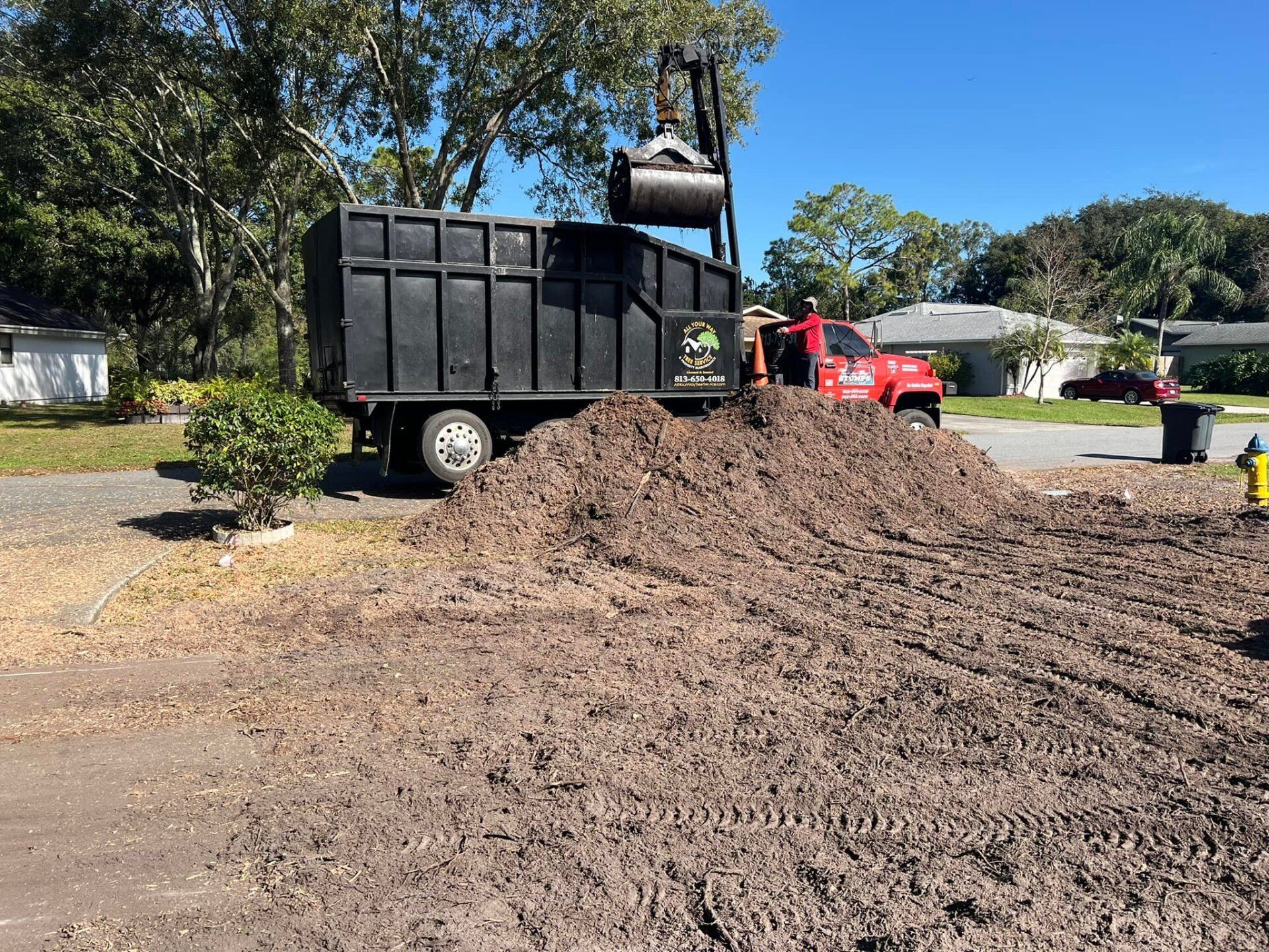 A truck loading mulch into a dumpster in a residential yard on a sunny day.