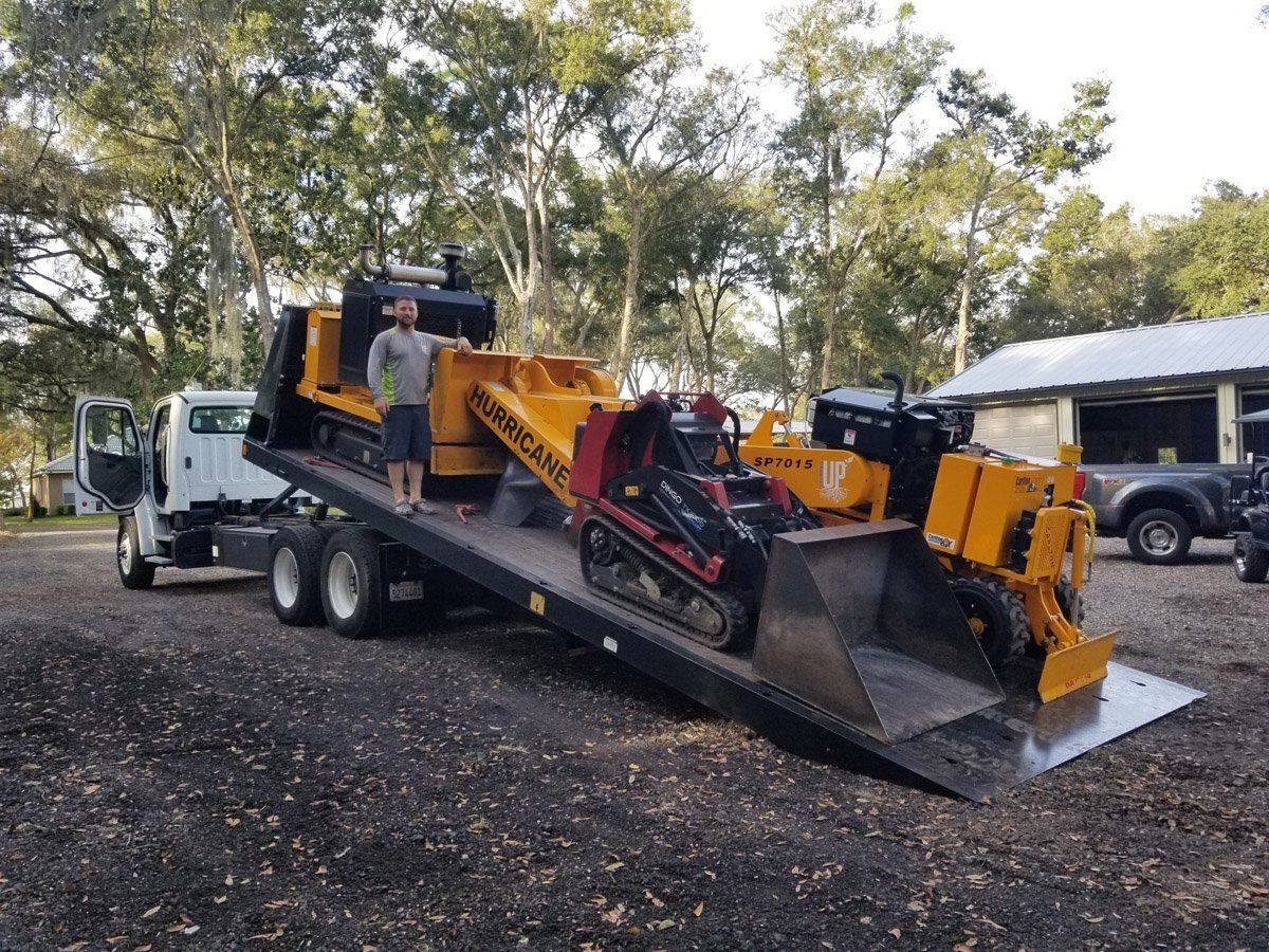 Man on flatbed truck with tree removal equipment: stump grinders and a mini skid steer. Outdoors.