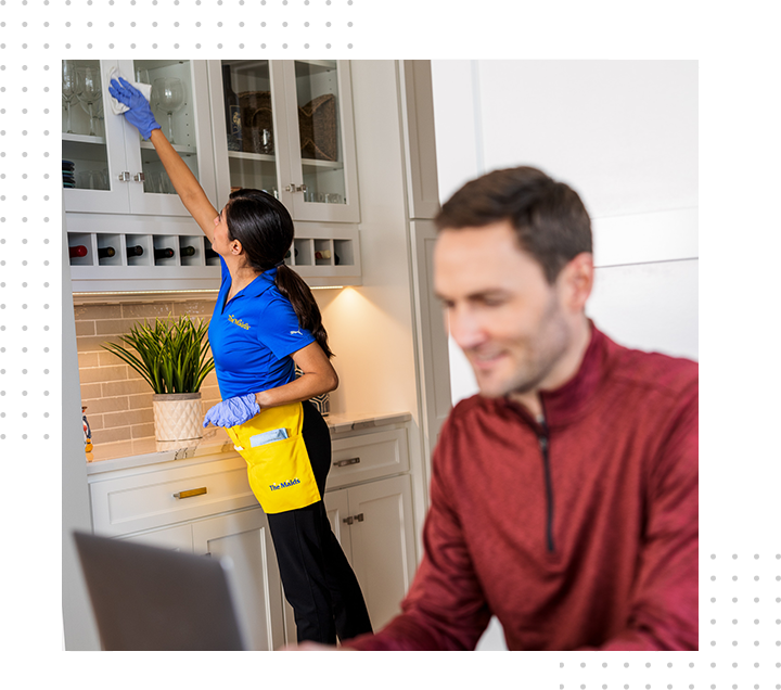 House cleaner in blue and yellow cleaning cabinets while a man sits blurred in the foreground