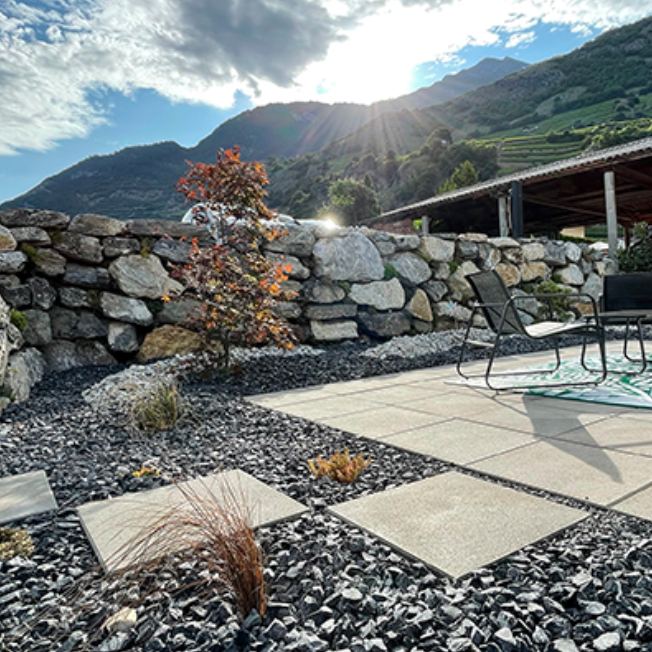 Un patio avec des chaises et une table devant un mur de pierre avec des montagnes en arrière-plan.