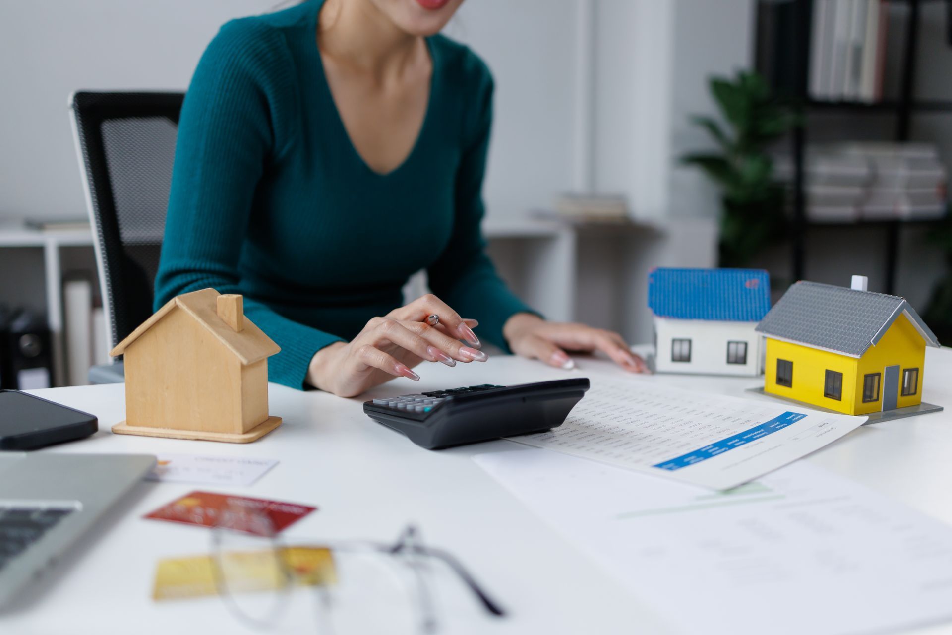 A cropped view of a woman sitting at a desk with miniature houses, a calculator, and documents.