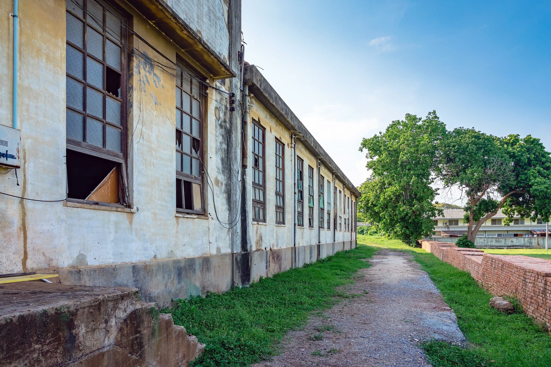 Old industrial building with broken windows beside a grassy path and trees in sunlight