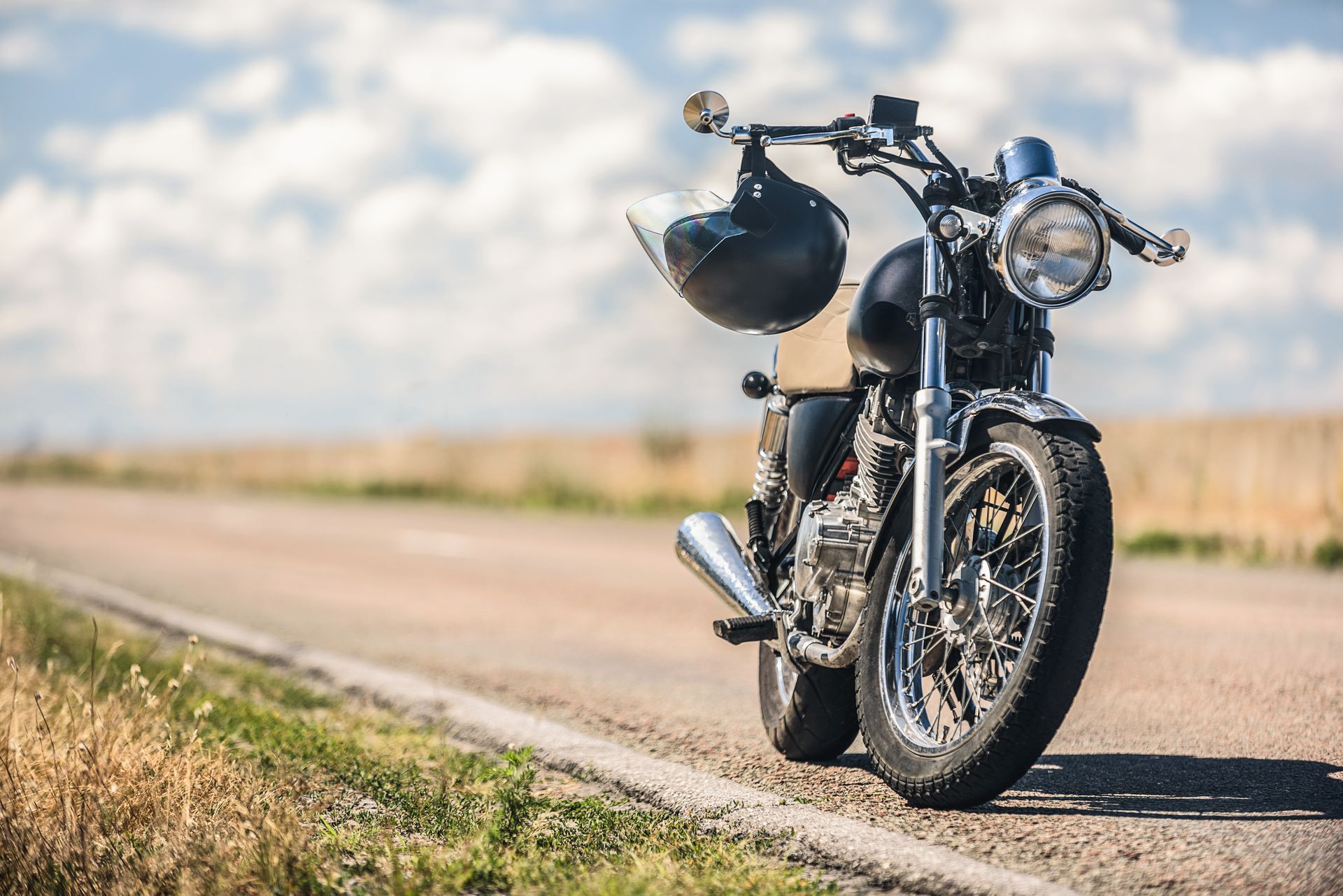 Black motorcycle parked on the side of a rural road, helmet hanging from the handlebars under a blue and cloudy sky.