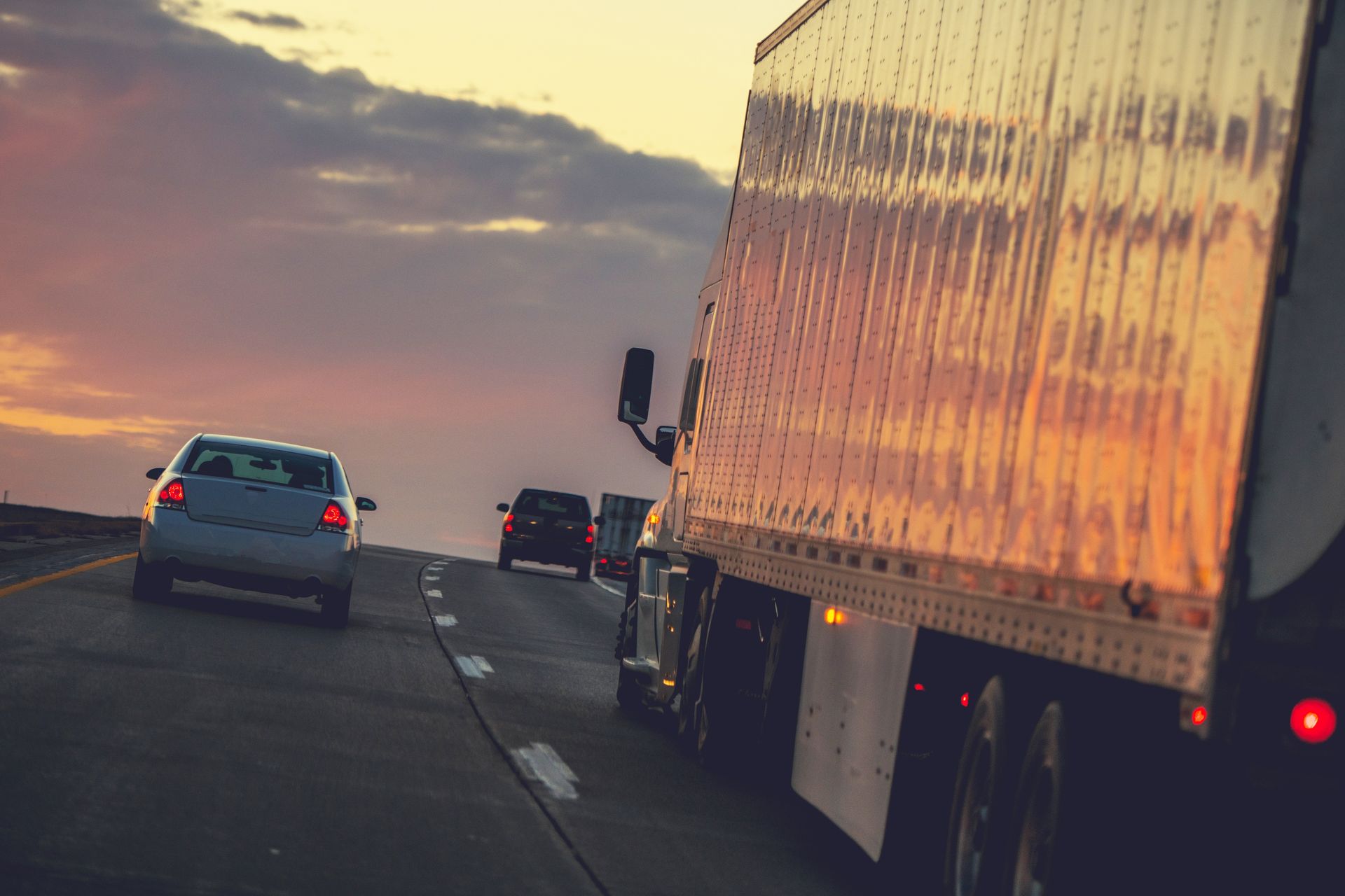 Semi-truck and cars on a highway at sunset, orange and purple sky reflecting on the truck.