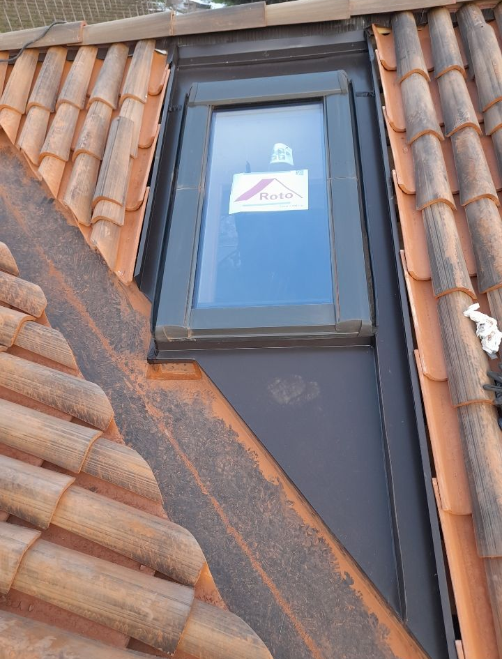 Finestra per tetto con cornice scura incastonata tra tegole di terracotta arancione su un tetto a forte pendenza.