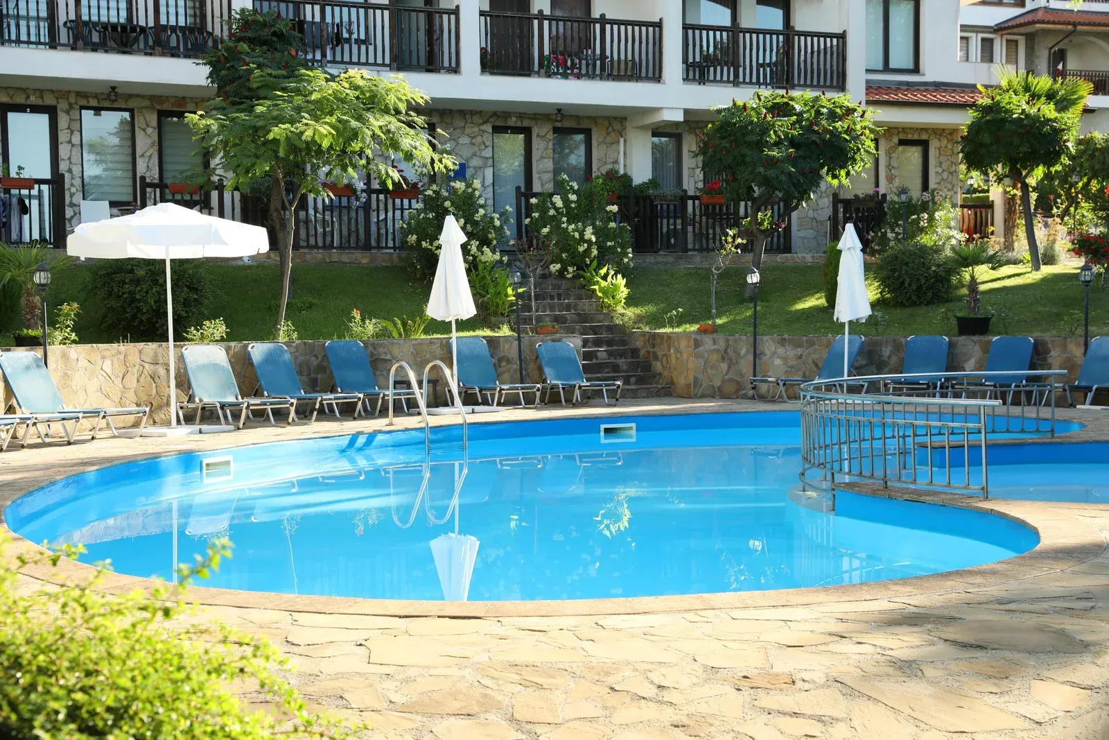 An outdoor swimming pool with bright blue water surrounded by lounge chairs, umbrellas, and a resort building.